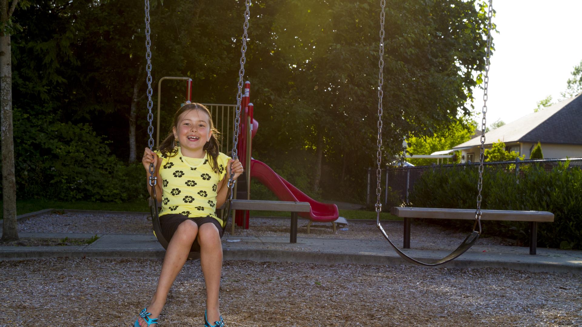 A young girl smiling on a swing. Behind her there are benches and a slide. 